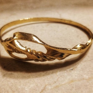 Puede incluir: Un brazalete de oro con un diseño abstracto retorcido. El brazalete está hecho de un metal liso y brillante.