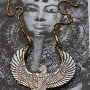 May include: A gold chain necklace with a large iridescent shell pendant depicting a winged goddess figure. The pendant is intricately carved with details of the goddess's wings, body, and face.
