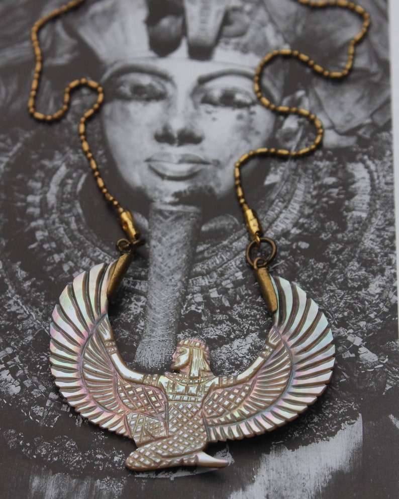 May include: A gold chain necklace with a large iridescent shell pendant depicting a winged goddess figure. The pendant is intricately carved with details of the goddess's wings, body, and face.