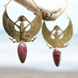 Puede incluir: Pendientes de aro dorados con un diseño de diosa alada y una piedra rosa colgante.