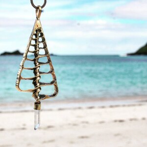 Puede incluir: Un collar con colgante dorado con un diseño calado único. El colgante tiene forma triangular con un detalle en espiral y un cristal transparente en la parte inferior. El collar cuelga sobre un fondo borroso de playa y océano.