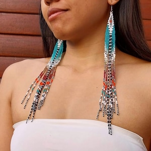 May include: Long, beaded earrings with a fringe design. The earrings feature a color palette of turquoise, white, red, black, and gold beads. The earrings are designed with a long, dangling style.