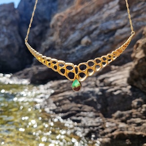 Op de afbeelding: Een gouden ketting met een uniek ontwerp met een reeks met elkaar verbonden cirkels. De ketting heeft een traanvormige hanger met een groene en bruine steen.