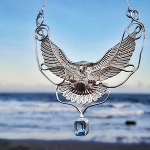 Puede incluir: Un collar de plata con un diseño de águila detallado y un colgante de piedra azul. El collar está fotografiado sobre un fondo borroso de playa y océano.