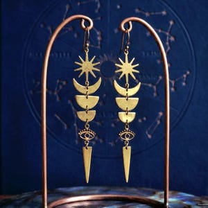 Puede incluir: Pendientes celestiales dorados exhibidos en un soporte de color cobre. Cada pendiente presenta un sol, medias lunas, un ojo y un colgante puntiagudo. El fondo es azul oscuro con un diseño de constelación.