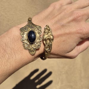 May include: Ornate brass cuff bracelet with a dark blue oval stone and a detailed, sculpted design. The bracelet features an open design with intricate patterns and a decorative clasp. The jewelry is displayed on a neutral background.