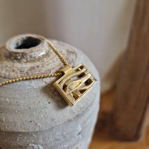 May include: Gold necklace with a rectangular pendant featuring an intricate design of an eye with a winged symbol. The pendant is on a chain.