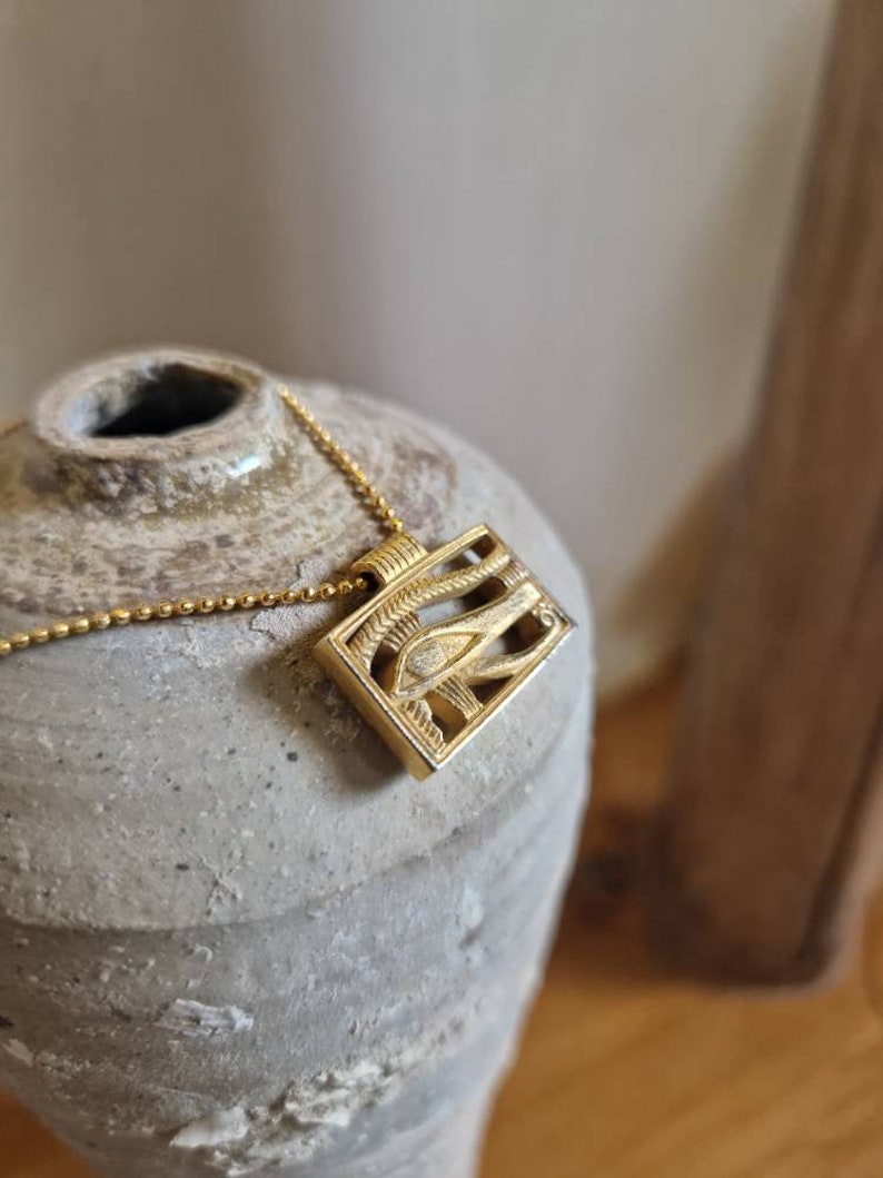 May include: Gold necklace with a rectangular pendant featuring an intricate design of an eye with a winged symbol. The pendant is on a chain.