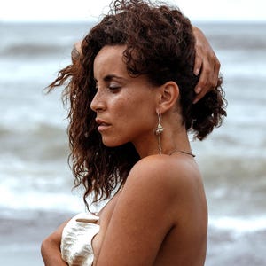 Puede incluir: Una mujer con cabello castaño rizado está de pie en una playa, sosteniendo una gran concha blanca. Lleva pendientes de concha plateados. El fondo muestra el océano con olas. Un tatuaje es visible en su costado.