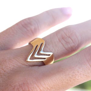 Puede incluir: Un anillo dorado con un diseño geométrico que presenta una serie de líneas anguladas. El anillo se lleva en un dedo, mostrando su forma única y brillo metálico. El diseño del anillo es moderno y llamativo.