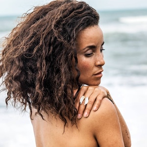 Puede incluir: Una mujer con cabello castaño rizado está de pie en una playa. Lleva un anillo con un diseño de ala blanca en su dedo. El océano está en el fondo.