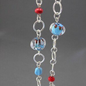 May include: A silver chain necklace with blue and red glass beads. The beads have a swirl pattern and are clear with a blue and red color.
