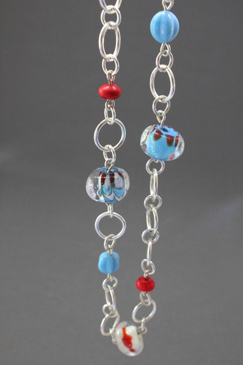 May include: A silver chain necklace with blue and red glass beads. The beads have a swirl pattern and are clear with a blue and red color.