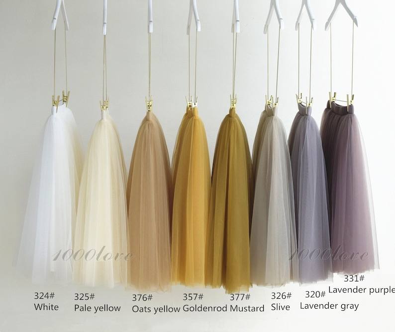 May include: Eight tulle skirts hanging on hangers, showcasing a variety of colors, including white, pale yellow, oats yellow, goldenrod, mustard, silver, lavender gray, and lavender purple. The skirts are all made of soft, flowing tulle and are perfect for adding a touch of elegance to any outfit.