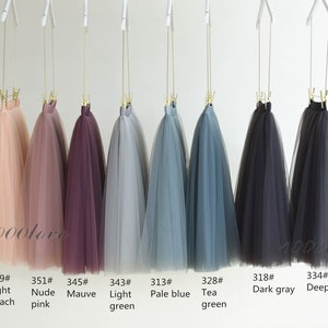 May include: Eight tulle skirts hanging on hangers, displayed in a row. The skirts are in various colors, including light peach, nude pink, mauve, light green, pale blue, tea green, dark gray, and deep gray. The skirts are labeled with numbers and color names.