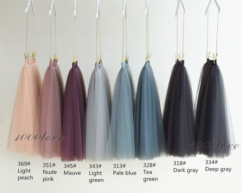 May include: Eight tulle skirts hanging on hangers, displayed in a row. The skirts are in various colors, including light peach, nude pink, mauve, light green, pale blue, tea green, dark gray, and deep gray. The skirts are labeled with numbers and color names.