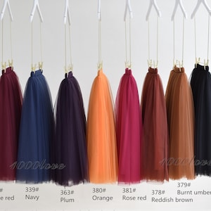 May include: Ten tulle skirts hanging on hangers. The skirts are in the following colors: wine red, navy, plum, orange, rose red, reddish brown, burnt umber, and black. Each skirt has a number tag attached to it.