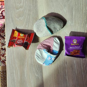 Puede incluir: Una roca plana de fieltro gris, una zapatilla de tela de patchwork rosa y azul, una bolsa morada de galletas Annie's Berry Patch y un paquete rojo y blanco de mini snacks Sun-Maid.