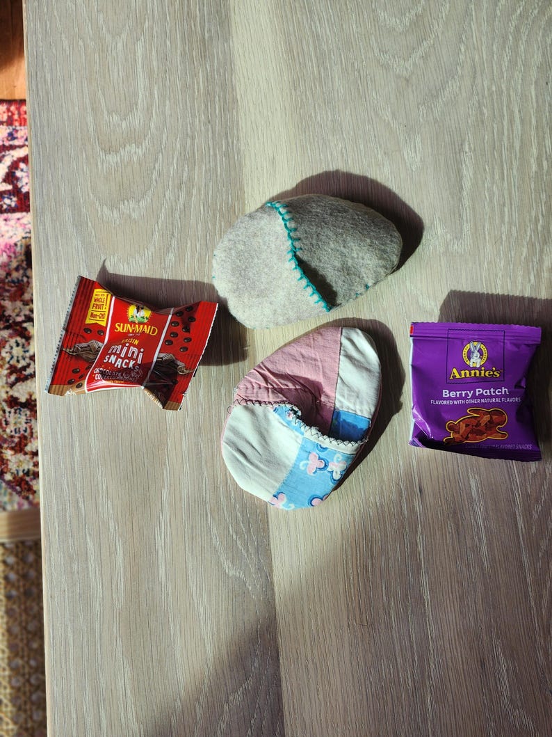 Puede incluir: Una roca plana de fieltro gris, una zapatilla de tela de patchwork rosa y azul, una bolsa morada de galletas Annie's Berry Patch y un paquete rojo y blanco de mini snacks Sun-Maid.