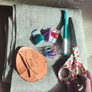 Puede incluir: Un proyecto artesanal con fieltro beige, un patr&oacute;n para un proyecto de fieltro, hilo de bordar verde, morado y rojo, un par de tijeras con estampado de fresa y un bol&iacute;grafo negro y blanco.