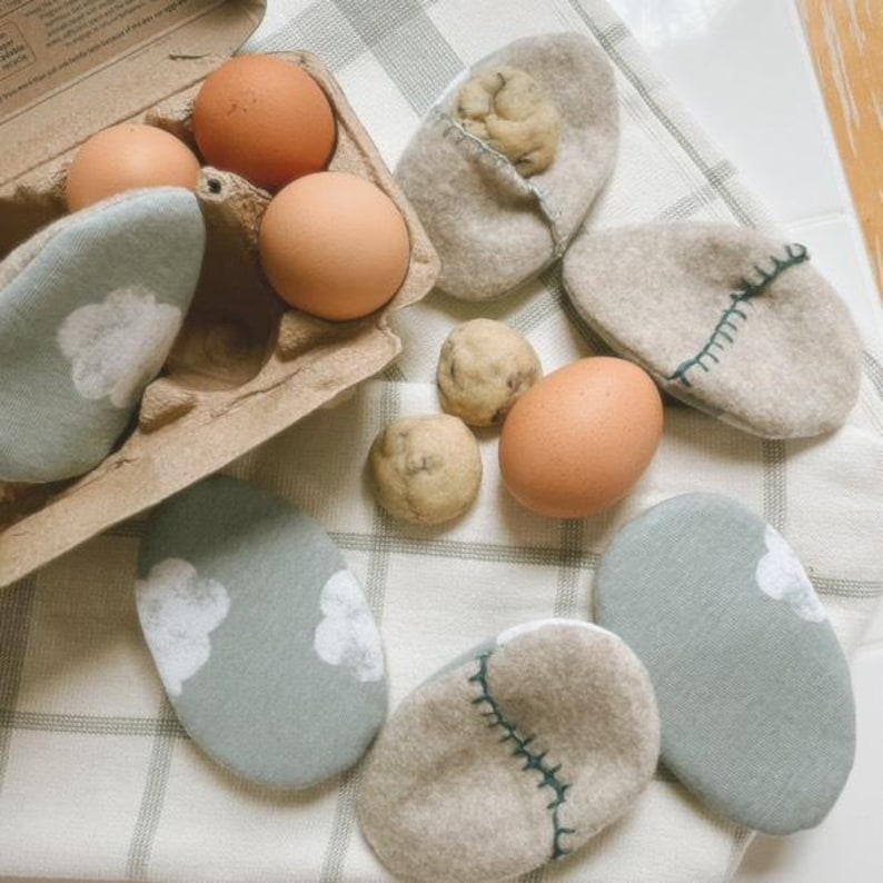 Puede incluir: Seis huevos marrones en una caja de cart&oacute;n para huevos, junto a seis bolsas de tela con forma de huevo. Tres bolsas son beige con costuras verdes, y tres bolsas son verdes con dise&ntilde;os de nubes blancas. Tambi&eacute;n hay tres galletas de chocolate con chispas marrones sobre una superficie a cuadros blancos y verdes.