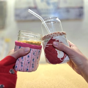 Może przedstawiać: Dwa szklane słoiki typu Mason z materiałowymi osłonami. Jeden słoik ma przezroczystą słomkę, a drugi zawiera żółty płyn. Osłony mają wzory kwiatowe i czerwone. Słoiki są trzymane w dłoniach.