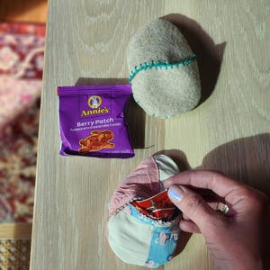Puede incluir: Un paquete de aperitivos Annie's Berry Patch morado con un logotipo de conejo y el texto "Berry Patch" y "Flavored with other natural flavors". Una mano sostiene una peque&ntilde;a bolsa de tela de retazos con un paquete de aperitivos rojo dentro.