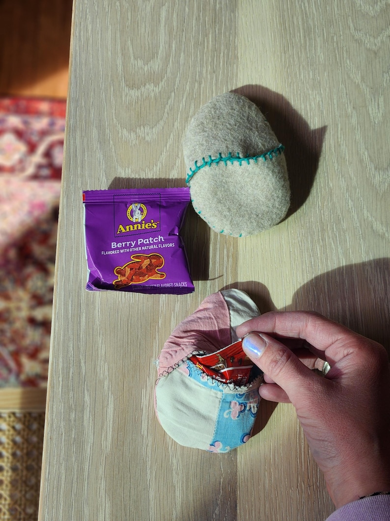 Puede incluir: Un paquete de aperitivos Annie's Berry Patch morado con un logotipo de conejo y el texto "Berry Patch" y "Flavored with other natural flavors". Una mano sostiene una peque&ntilde;a bolsa de tela de retazos con un paquete de aperitivos rojo dentro.