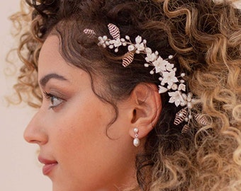 May include: A woman with curly hair wears a white lace dress and a floral hairpiece with pearl accents. The hairpiece is made of delicate white flowers and gold leaves.