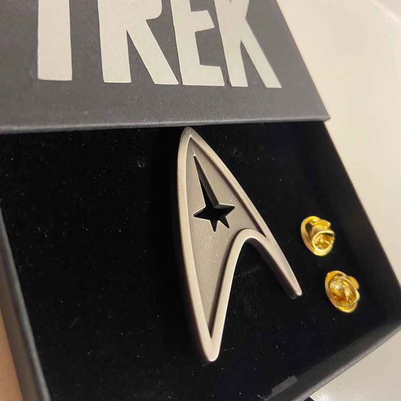 May include: A silver Starfleet insignia pin in a black velvet box with the word "TREK" printed on the lid. The pin is a stylized representation of the Starfleet insignia, with a star in the center and a pointed shape.