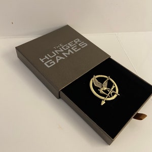 May include: A gold-colored pin featuring the Mockingjay symbol from The Hunger Games. The pin is in a gray box with the words "The Hunger Games" printed on the top.