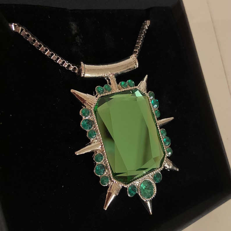 Puede incluir: Collar plateado con un gran colgante rectangular de piedra verde. El colgante est&aacute; enmarcado por peque&ntilde;as gemas verdes y puntas plateadas. El collar se presenta sobre una superficie negra.