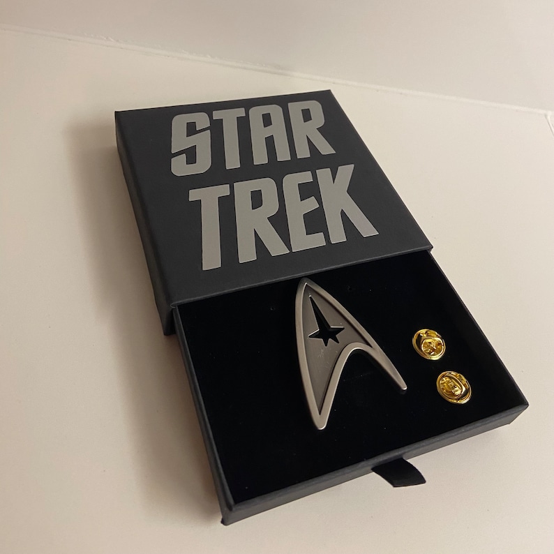 May include: A black box with the words "STAR TREK" printed in silver on the top. The box is open and contains a silver Star Trek insignia pin and two gold pins.