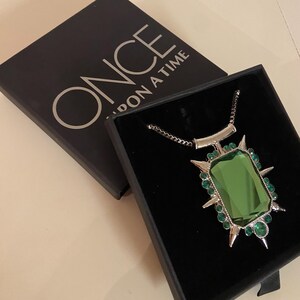 Puede incluir: Un collar plateado con un gran colgante rectangular de piedra verde. El colgante est&aacute; rodeado de peque&ntilde;as gemas verdes y puntas plateadas. El collar se presenta en una caja negra con la inscripci&oacute;n "ONCE UPON A TIME" en la tapa.