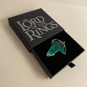 May include: A black box with the words "The Lord of the Rings" printed on the top. The box is open and a silver and green leaf pin is inside.