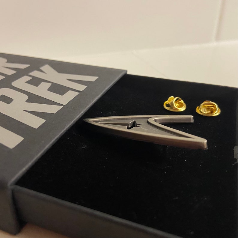 May include: A silver Star Trek insignia pin in a black box with the word "TREK" printed on the lid. The pin is in the shape of a starfleet delta with a star in the center. Two gold pins are visible on the black velvet lining of the box.