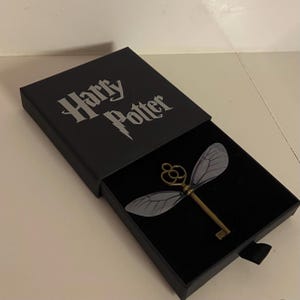 May include: A black box with the words "Harry Potter" in silver lettering. Inside, a bronze-colored key with delicate, translucent wings is displayed against a black velvet background. The key is a decorative item.