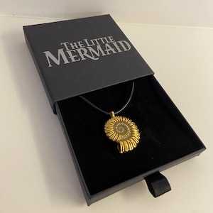 Peut inclure: Un pendentif doré en forme de coquillage en spirale, présenté dans un coffret noir ouvert. Le coffret porte l'inscription "THE LITTLE MERMAID" en lettres argentées. Le pendentif est suspendu à un cordon noir.