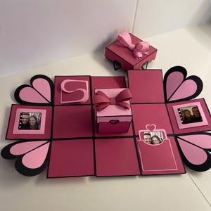 Op de afbeelding: Een roze en zwarte explosiebox met een hartvormig ontwerp. De doos heeft meerdere compartimenten die opengaan om foto's en een kleine roze doos met een strik te onthullen.