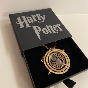 May include: A gold-colored necklace with a round pendant featuring a spinning hourglass design. The pendant is inscribed with stars and the necklace is in a black box with the words "Harry Potter" printed on the top.
