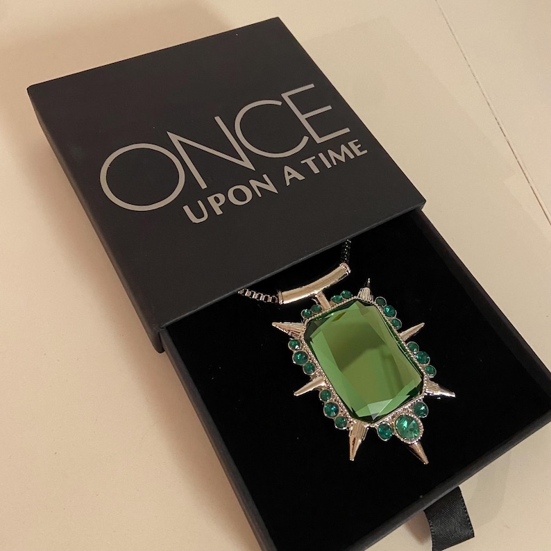 Puede incluir: Un collar con colgante de piedra preciosa verde en una caja negra. El colgante tiene una gran piedra verde rectangular rodeada de piedras verdes m&aacute;s peque&ntilde;as y detalles plateados. La caja est&aacute; abierta, revelando el collar, y tiene las palabras "ONCE UPON A TIME" impresas en la tapa.