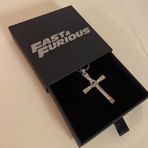 Può includere: Un ciondolo a forma di croce argentata con cristalli trasparenti, esposto in una scatola nera aperta. La scatola ha le parole "FAST & FURIOUS" in lettere argentate sul coperchio. La collana è su una catena d'argento.