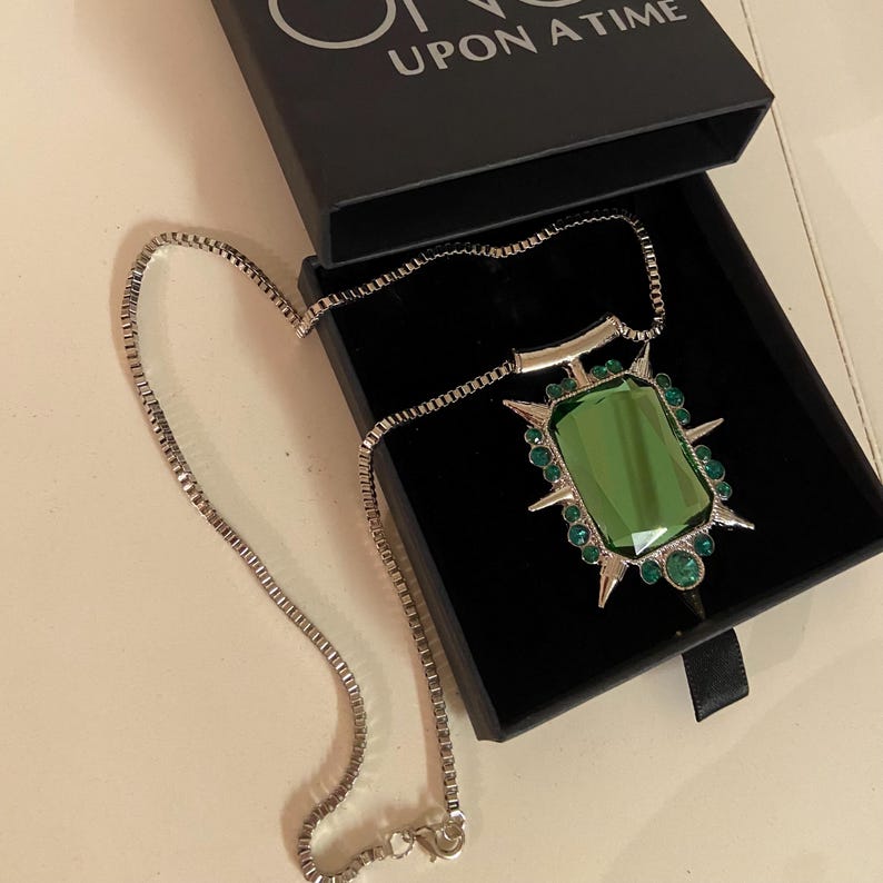 Puede incluir: Un collar plateado con un gran colgante rectangular de piedra verde. El colgante est&aacute; rodeado de piedras verdes m&aacute;s peque&ntilde;as y puntas plateadas. El collar se presenta en una caja negra con la inscripci&oacute;n "ONCE UPON A TIME" en la tapa.