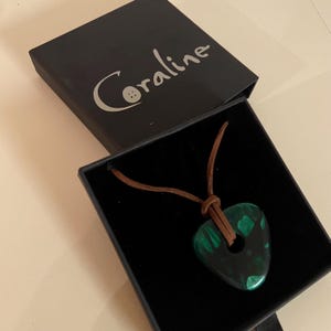 Può includere: Una collana con pendente verde e nero con cordino in pelle, presentata in una scatola nera. La scatola è aperta, rivelando la collana con una forma organica unica. La scatola è etichettata "Coraline" in un carattere scritto a mano.