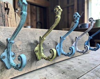 May include: A wall-mounted coat rack featuring five ornate metal hooks in shades of turquoise, yellow, light blue, white, and dark blue, all with a distressed finish, attached to a horizontal piece of weathered, rough-textured natural wood.