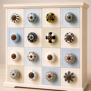 Op de afbeelding: Een crèmekleurige houten opbergkast met zestien vierkante lades, elk voorzien van een unieke, ronde, decoratieve keramische knop. De knoppen tonen diverse patronen, waaronder bloemmotieven, geometrische vormen en strepen in blauw, zwart, wit, geel en bruin, afgewerkt met een centraal bronzen metalen accent.