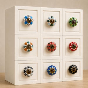 Può includere: Un mobiletto bianco con nove cassetti quadrati, ognuno dotato di un pomello in ceramica rotondo dipinto a mano. I pomelli presentano vari motivi floreali e geometrici in colori tra cui blu, verde, arancione, rosso, nero e bianco.