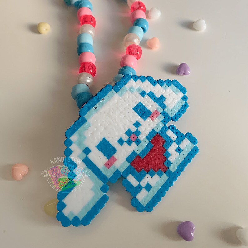 May include: A blue and white pixelated charm with a red heart in the center, hanging from a multi-colored beaded necklace.