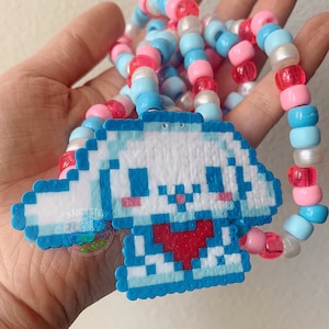 May include: A blue and white pixelated charm of a cartoon character with a red heart, attached to a beaded necklace with pink, blue, and white beads.