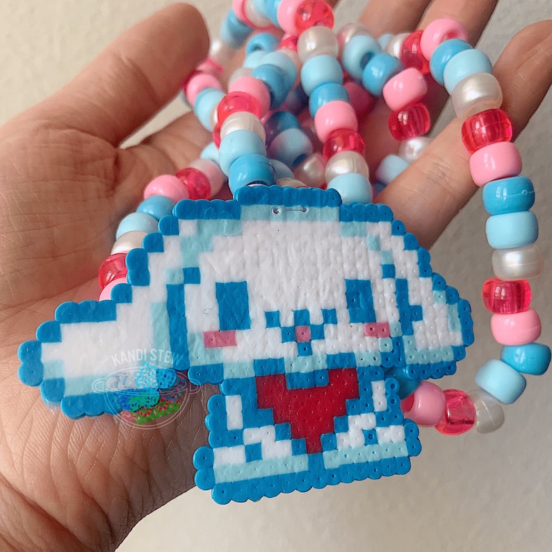 May include: A blue and white pixelated charm of a cartoon character with a red heart, attached to a beaded necklace with pink, blue, and white beads.
